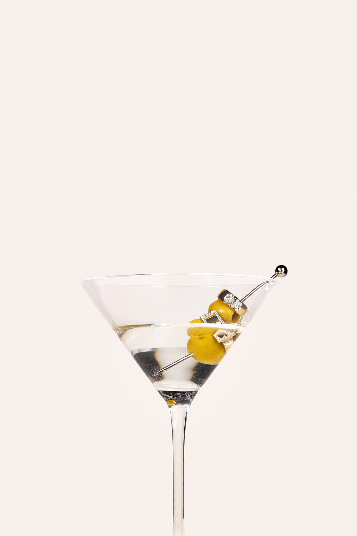 A martini glass with a silver cocktail stick, featuring two olives and two gold rings.