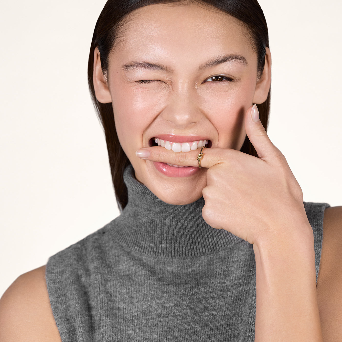 A young woman winking with one eye and putting her index finger between her teeth, while wearing the 14K gold Karli Knot Ring