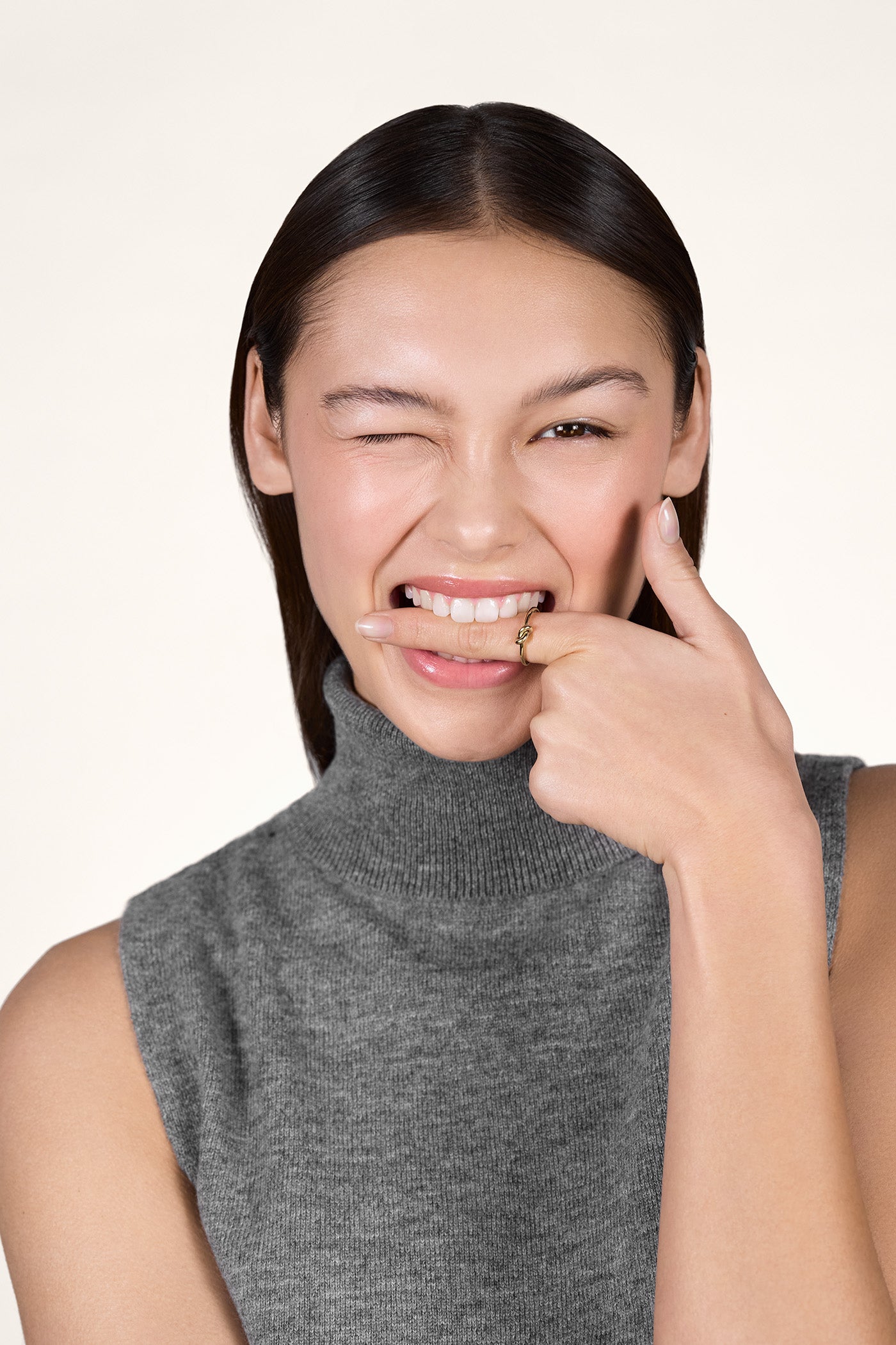 A young woman winking with one eye and putting her index finger between her teeth, while wearing the 14K gold Karli Knot Ring