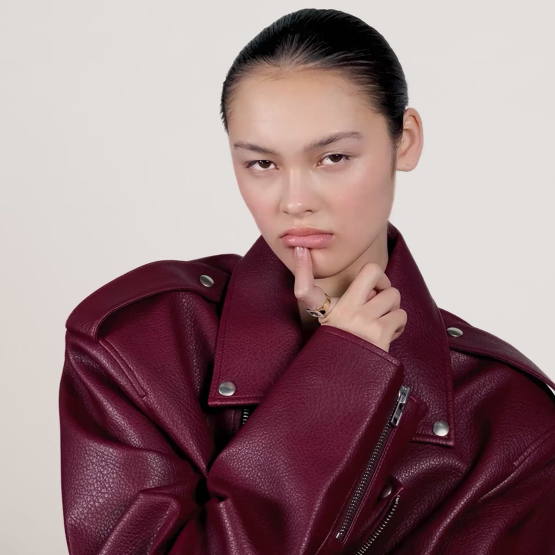 A video of a young woman wearing a burgundy red leather jacket and wearing a gold ring on her index finger.